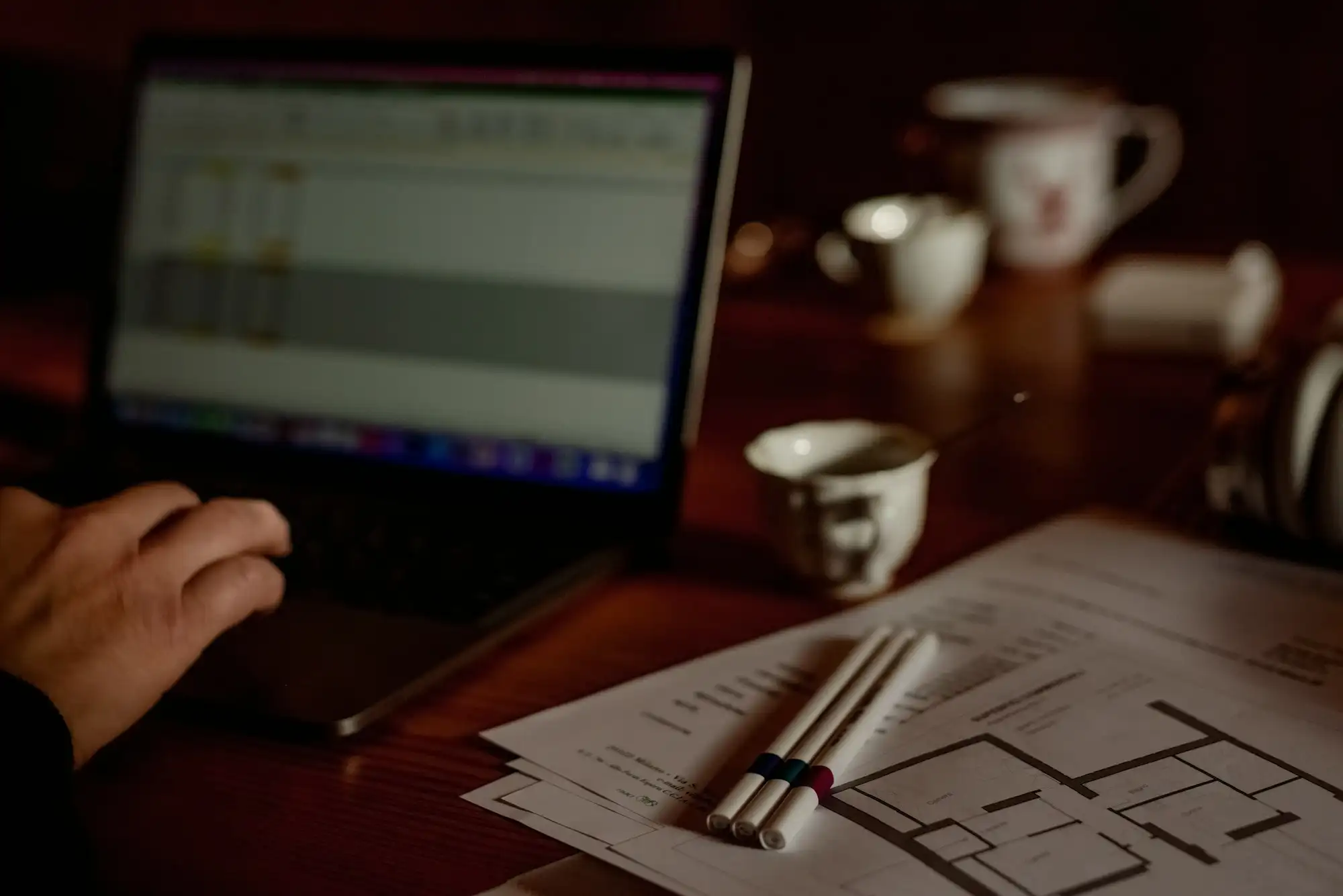 Main sur le clavier d’un ordinateur portable affichant un tableur, à côté de plans d’étage imprimés.